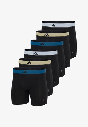 Junger Mann steht barfuß und trägt schwarze Boxer mit weißem Bund und Adidas-Logo vor weißem Hintergrund.