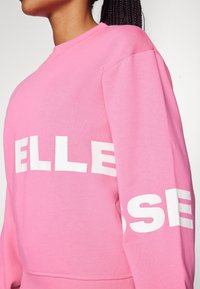 Sweat-shirt rose avec impression blanche. Présente un col rond et des manches longues, avec de grandes lettres sur la manche. Fabriqué en coton doux.