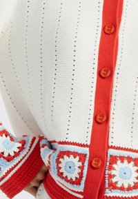  Gebreide cardigan in het wit met rode knopen en accenten. Bevat gehaakte bloempatronen in rood, wit en blauw bij de mouwen.