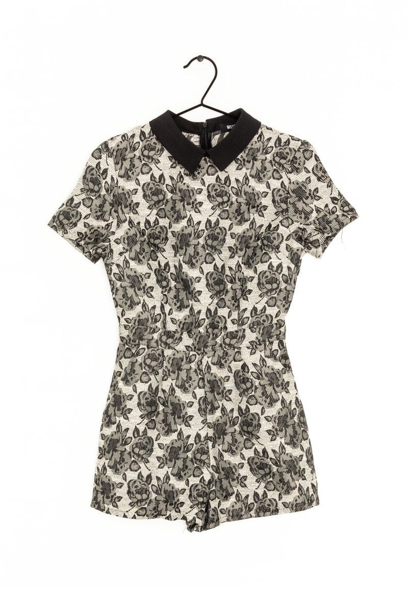 Barboteuse à manches courtes avec motif floral noir sur tissu blanc, avec un col Peter Pan noir, suspendue sur un cintre noir.