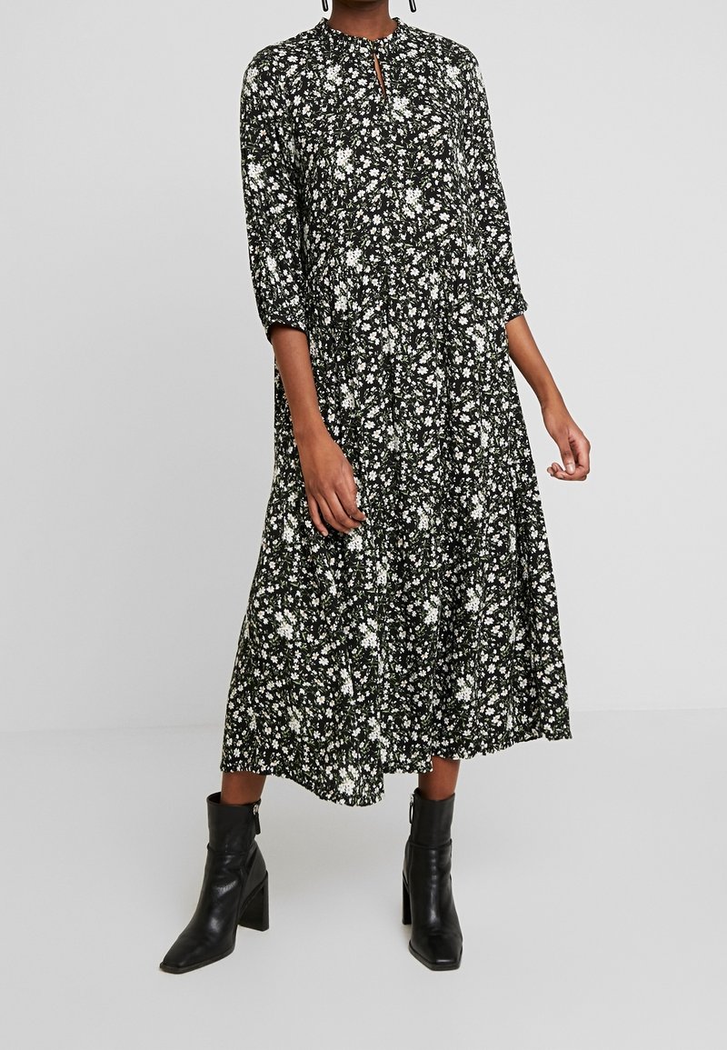 Personne portant une robe midi noire à motif floral blanc et manches trois-quarts, associée à des bottines noires à talons.