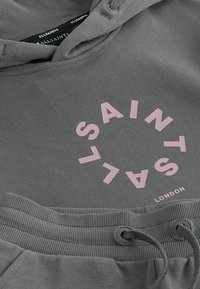 Sivá mikina AllSaints s kapucňou a tepláky s ružovým kruhovým logom a detailom sťahovacieho pásu na šnúrku.