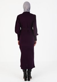 Langes, figurbetontes lila Kleid mit gepufften Ärmeln, tailliertem Gürtel und knielanger Länge, kombiniert mit knielangen schwarzen Stiefeln mit Absatz.
