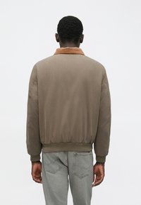 Άνδρας στέκεται με την πλάτη στραμμένη, φορώντας ένα καφέ bomber μπουφάν με κόκκο κλωστής και ανοιχτό γκρι τζιν μπροστά σε λευκό φόντο.