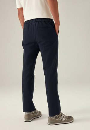 Marineblaue Hose mit elastischem Bund und geradem Schnitt, ausgestattet mit Gesäßtaschen und kombiniert mit grauen Sportschuhen. Glatte Stofftextur.