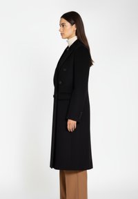 Sort dobbeltradet frakke med revers, struktureret stof og forlommer, parret med en beige turtleneck og skræddersyede brune bukser.