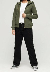 Sudadera verde oliva con cremallera y capucha, puños acanalados y bolsillos laterales, combinada con pantalones negros de pierna recta y zapatillas negras.
