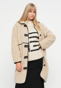 Beige γούνινο παλτό με γιακά, πέντε κουμπιά και αντίθετη καφέ περίθαλψη. Φοριέται πάνω από ένα κρεμ πουλόβερ με μαύρα γεωμετρικά σχέδια.