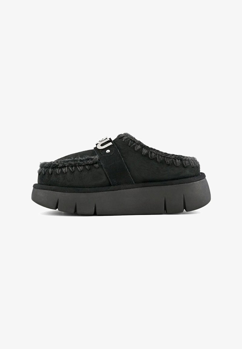 Musta mokkanahka slip-on -kenkä, jossa on turkisreunus, punottu ommel ja paksu musta pohja. Yläosassa on merkkistrappi.