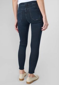 Jean skinny bleu foncé avec une texture lisse, deux poches arrière et une taille mi-haute, associés à des chaussures slip-on de couleur claire.