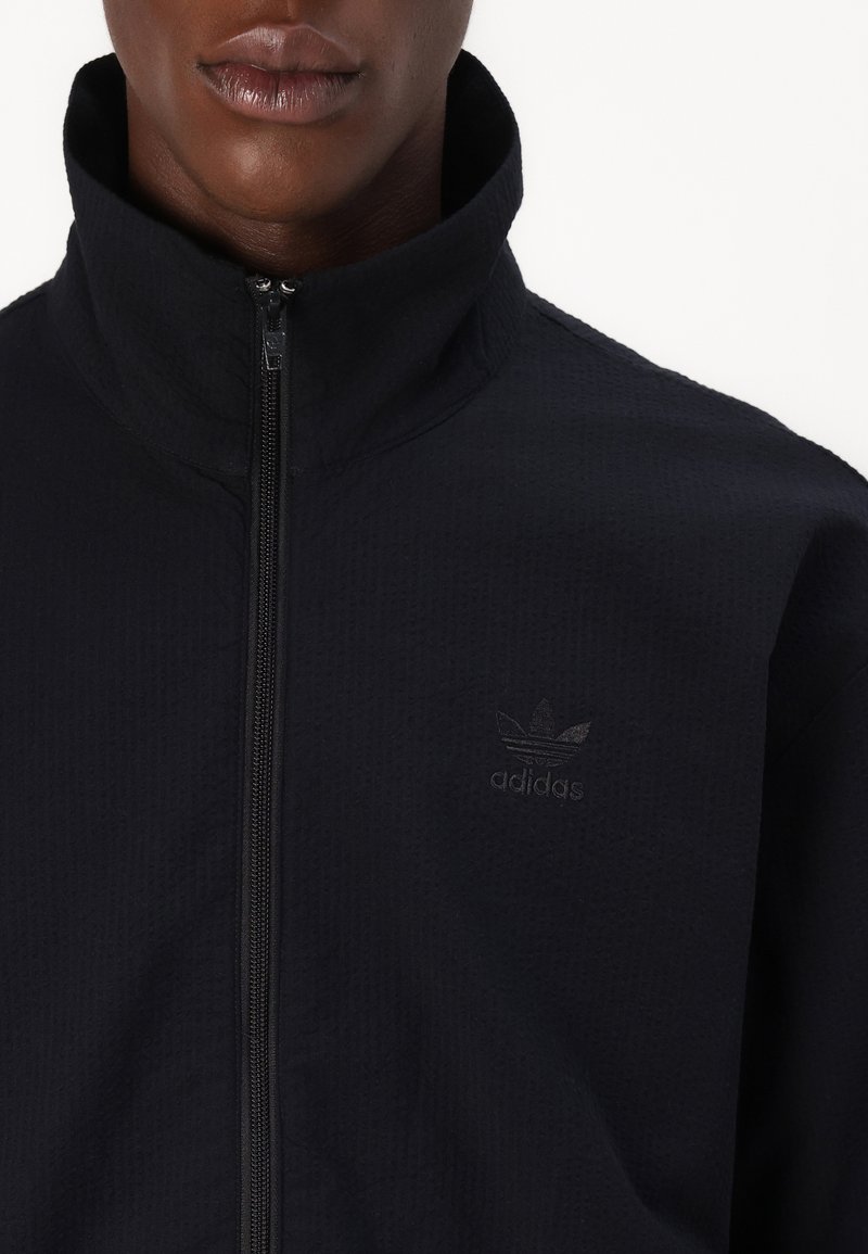 Schwarze Zip-Jacke mit hohem Kragen und strukturiertem Stoff, mit einem dezenten schwarzen adidas-Logo auf der Brust.