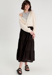 Cárdigan oversized beige claro, camiseta gris sin mangas, falda maxi negra de volantes y sandalias negras con tachuelas. Textura acogedora y diseño casual.