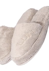 Beige tofflor gjorda av mjukt, plyschigt tyg med stängt tåparti, med en sömdetalj och en präglad logotyp på ovansidan.