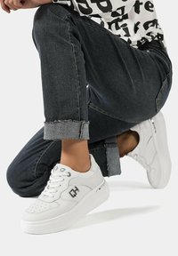 Sneaker bianco con punta perforata, lacci piatti e pannelli testurizzati. Presenta suola spessa bianca e dettagli metallici. Indossato con jeans scuri con risvolto.