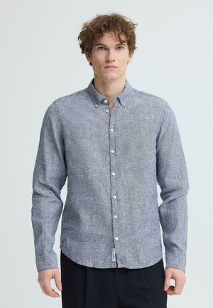 Młody mężczyzna z kręconymi włosami, ubrany w jasnoszarą koszulę z długim rękawem zapinaną na guziki oraz ciemne spodnie, stojący na tle jednolitego jasnego tła.