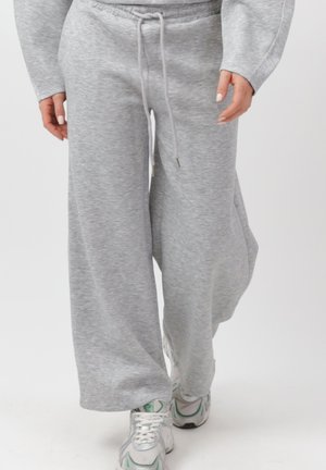 Osoba ubrana w luźne, jasnoszare spodnie dresowe z sznurkiem i białe trampki, idąca na tle jednolitego tła.