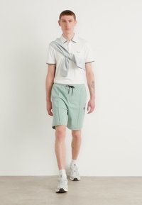 Tenue masculine comprenant un polo blanc avec des accents verts, un short vert clair avec un cordon de serrage et des baskets blanches. Design simple, style décontracté.