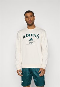 Sweatshirt crème avec "ADIDAS TENNIS CLUB" imprimé en vert foncé. Assorti avec un short teal arborant un logo blanc. Texture de tissu lisse.