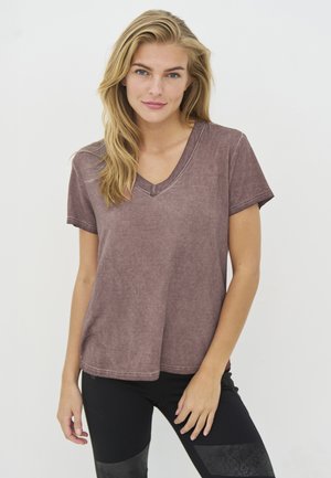 Jonge vrouw met lang blond haar die een uitgebleekt paarse V-hals t-shirt met korte mouwen en een zwarte broek draagt, staande tegen een effen witte achtergrond.