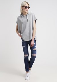 Felpa grigia con cappuccio e lacci, abbinata a jeans scuri strappati e sneakers bianche. Il modello indossa occhiali da sole neri.