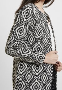 Cardigan a motivi bianco e nero con un design a diamante, realizzato in tessuto testurizzato, con apertura frontale e maniche lunghe.