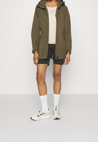 Person wearing olive green jacket, beige top, black shorts, white crew socks, and beige sneakers standing on gray floor.