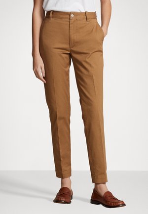 Woman wearing slim-fit brown chinos with side pockets paired with brown leather loafers, standing against a white background.