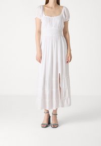 Vit maxiklänning med kort puffärm, fyrkantig halsringning och resår i midjan. Har en slits framtill och en vågig nedre del. Matchas med blå klacksandaler.