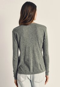 Kvinna med långt brunt hår som bär en åtsittande grå långärmad tröja och ljusblå jeans, vänd bort från kameran mot en enkel bakgrund.