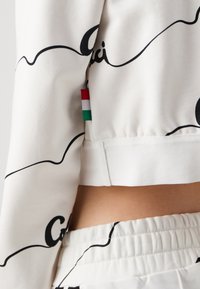 Gros plan sur un sweat-shirt blanc et un pantalon assorti avec un motif de lignes abstraites noires et une petite étiquette de drapeau rouge, blanc et vert sur la couture latérale.