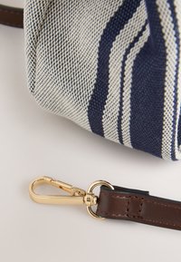 Détail d'un sac tissé beige et bleu marine rayé avec un fermoir en or et une bandoulière en cuir marron sur fond blanc.