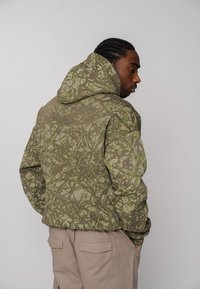 Veste à capuche au motif camouflage vert et marron, avec une coupe décontractée et un tissu texturé. Vue de dos mettant en valeur le design et les détails de couture.