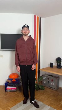 Bordo dukserica s patentnim zatvaračem i natpisom sprijeda, crne hlače i smeđe cipele. Samostalna figura na minimalno uređenoj unutarnjoj pozadini.