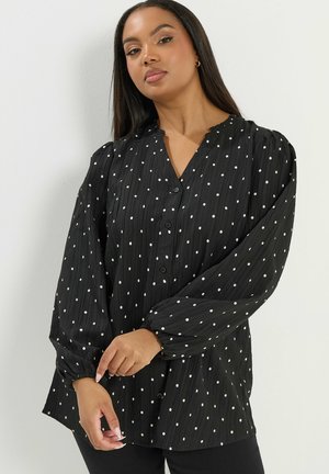 Kvinde iført en sort, langærmet, knap-op bluse med hvide polkaprikker, som justerer manchet på venstre håndled.