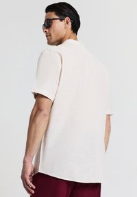 Uomo che indossa una camicia a maniche corte beige chiaro e occhiali da sole scuri, girato di spalle, con pantaloni bordeaux su uno sfondo semplice e chiaro.