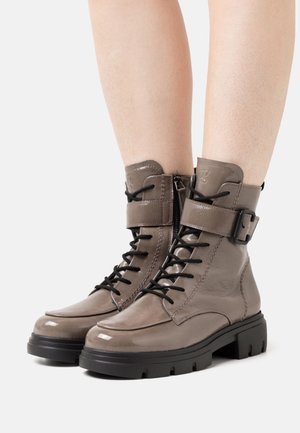 Bottes à lacets brillantes couleur taupe avec semelles épaisses noires, fermetures éclair latérales et sangles à boucle, portées sur des jambes nues sur un fond blanc.