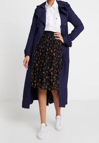 Kvinna iklädd en marinblå trenchcoat, vit skjorta med krage, svart blommig plisserad kjol och vita sneakers, stående med ena handen på höften.