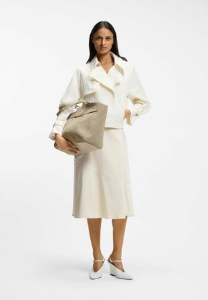 Kvinna i cremefärgad jacka och kjol som håller en stor beige handväska, bär vita högklackade skor mot en vit bakgrund.