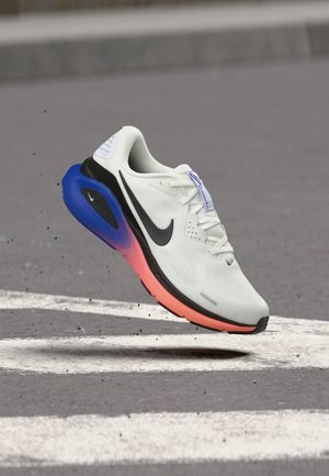 Hvid og sort Nike løbesko med blå og pink sål, der svæver over grå fortov med hvide vejmarkeringer.