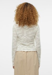 Langermet topp i lys kremfarge med en teksturert, gjennomsiktig design med varierende utskjæringer. Parret med et beige, lett skjørt.