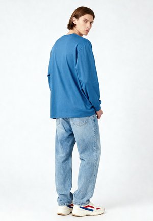 Jonge man met halflang haar die naar de camera draait, gekleed in een blauw shirt met lange mouwen, lichtgewassen jeans en witte sneakers met rode en blauwe details.