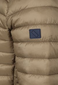 Beige pufferjas met een gequilt ontwerp, voorzien van een blauw rechthoekig logo-patch op de borst. Glad, nylonachtig textuur.