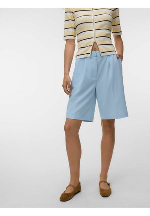 Femeie care poartă pantaloni scurți albastru deschis până la genunchi, tricou galben în dungi cu mâneci scurte și fermoar, și pantofi maro Mary Jane cu talpă joasă, stând cu mâna în buzunar.