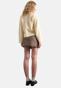 Femme aux cheveux blonds bouclés portant un pull écru, une mini-jupe à carreaux marron, des chaussettes beiges et des chaussures noires, vue de dos sur un fond blanc.