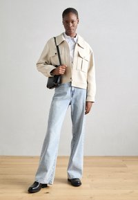 Giacca corta beige con grandi tasche, indossata sopra una camicia a righe, abbinata a jeans a gamba larga blu chiaro e stivaletti neri. Borsa a tracolla nera.