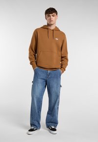 Un giovane uomo sta in piedi con sicurezza, indossando un hoodie marrone e jeans blu larghi, con le mani in tasca, su uno sfondo grigio uniforme.