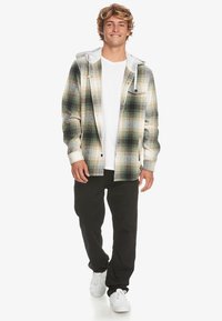 Ung man som går, iklädd en grön och beige rutjacka över en vit t-shirt, svarta byxor och vita sneakers, mot en vit bakgrund.