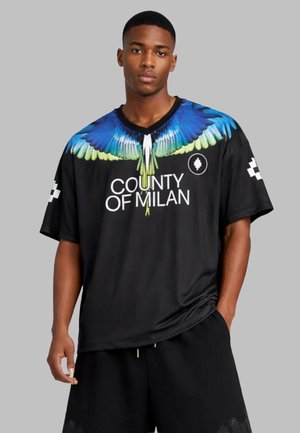 Junger Mann trägt schwarzes oversized T-Shirt mit blau-grünem Vogelflügel-Design auf den Schultern und dem Schriftzug "County of Milan" vorne.