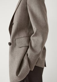 Vue latérale d'une personne portant un blazer gris avec des boutons et une poche, associé à un pantalon foncé sur un fond uni.