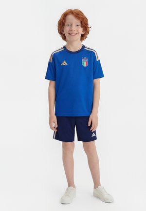 Usmievavý ryšavý chlapec stojaci, oblečený v modrom futbalovom drese Talianska, tmavomodré šortky s bielymi pruhmi a biele tenisky na bielom pozadí.